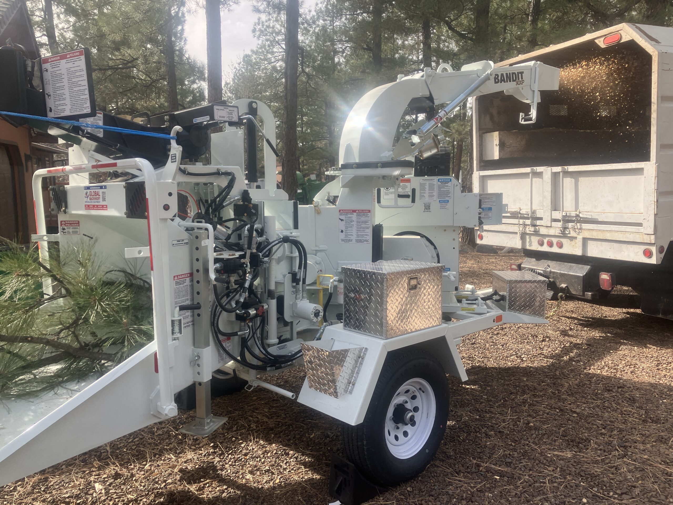 A wood chipper processes tree branches, ejecting wood chips into a truck parked nearby in an outdoor setting with trees in the background.